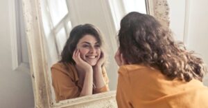 A cheerful woman smiles at her reflection in a vintage-style mirror, exuding positivity and warmth.